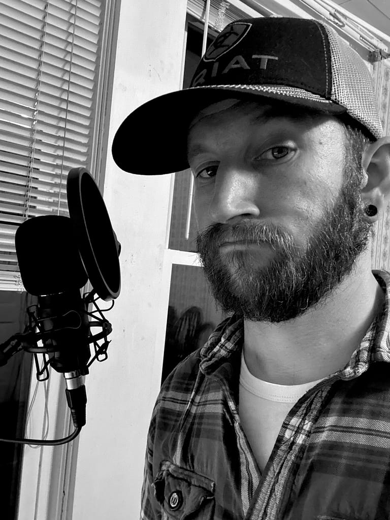 Black-and-white photo of Thomas Rocharz from The Irreverent Tenet, near a podcast microphone in a flannel shirt and baseball cap
